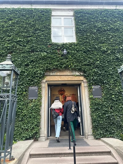 The image captures the main entrance to the Louisiana Museum of Modern Art in Humlebæk, Denmark. The building's facade is extensively covered in dense, dark green ivy. Centered above the entrance, a white-framed window with four panes is visible. A dark-colored security camera is mounted on the ivy-covered wall below the window and to the right of a small, recessed light fixture. The entrance itself features a prominent stone archway framing open double doors.
Two dark rectangular signs with white text are affixed to the ivy on either side of the entrance. The sign to the left indicates "KLUBKORT E-TICKETS" with a right arrow, and "LOUISIANA." The sign to the right indicates "BILLETTER TICKETS" with a left arrow, and "LOUISIANA."
Two individuals are depicted from behind, ascending two grey stone steps and entering the building through the open doors. The person on the left wears a red top, light blue jeans, a light-colored shawl or wrap over their shoulders, and carries a black crossbody bag. The person on the right wears a dark jacket, dark green trousers, and carries a light-colored shoulder bag with a patterned strap.
Through the open doors, a large, ornate copper-colored light fixture is visible hanging from the ceiling, along with posters on the interior wall that display "LOUISIANA" and "KAARI UPSON." To the far left, a decorative black metal lantern structure, partially obscured, shows an exposed lightbulb element. A black metal handrail extends from the bottom of the frame towards the entrance steps. The sky above the building is overcast.