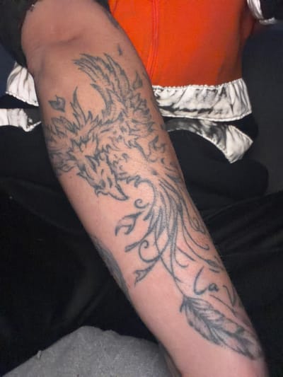 The image shows a person's forearm with a large tattoo of a phoenix with feathers and swirling patterns. The tattoo is in black ink. The person is wearing a gray shirt on the left and an orange garment with a white and black patterned item on the right. The background is dark and blurry. There is no information indicating this image was taken in Sant Martí Sarroca, Spain.