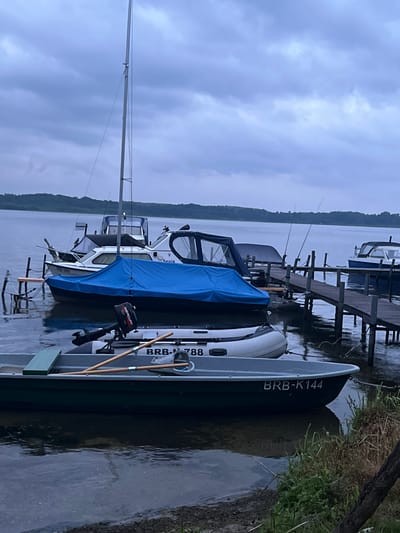 A cloudy, overcast day at a marina on Lake Schwielowsee, Germany. Several boats are docked. In the foreground, a dark green metal boat with the registration "BRB-K144" is moored closest to the shore. Behind it is a white inflatable boat with a small outboard motor and two wooden oars resting inside. A larger sailboat with a blue cover is docked behind the inflatable boat, and further back, a white cabin cruiser with the name "Sepia" is visible. A wooden pier extends from the shore to the right, with several fishing rods propped up. The lake stretches out to a tree-lined horizon in the background. The water is calm and reflects the muted sky. No people are present in the image.