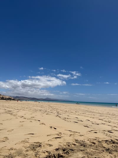 The image shows a beach in Pájara, Spain. The sand is a light brown color and covered in footprints. The sky is a clear blue with some white clouds. There is a body of water in the distance. There are a few people visible on the beach and in the water.