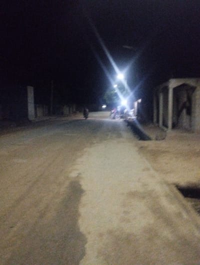 The image depicts a night scene in Bajoga, Nigeria, captured from a street level perspective. The scene is illuminated by a bright streetlight casting a strong glare. The road, with a dirt-like surface, stretches out from the viewer towards the light source, and is bordered by a concrete sidewalk to the right. Several people are congregated to the right of the road near a building, the details of which are blurred and difficult to discern. The scene is dominated by the bright streetlight, which casts a strong glare, making it difficult to see details beyond the immediate vicinity.