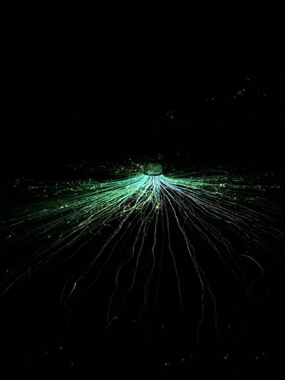 A long-exposure photograph depicts a dark, nocturnal environment identified as the city of Bukovik, Montenegro. A singular, luminous, roughly conical or bowl-shaped object, exhibiting a faint blue-green glow, is situated centrally in the upper mid-ground. From this object, and extending radially downwards and outwards, are numerous elongated, curvilinear light trails. These trails are predominantly green and light blue, with some fading into yellowish or white hues, and vary in thickness and intensity. They form intricate, winding patterns against the deep black background. The surrounding environment is characterized by this pervasive darkness, indicative of a night scene, with only a few minute, faint points of light sparsely visible in the far background and upper edges of the frame. No human figures, animals, or specific interactions between subjects are discernible; the scene focuses solely on the static glowing object and the dynamic trajectories of the light trails.