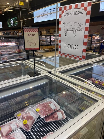 The image shows a refrigerated display case filled with packaged pork cuts in a supermarket in Saint-Brice-Courcelles, France (coordinates: 49.283, 3.999). Above the case, a sign reads "BOUCHERIE" with a cartoon pig and the word "PORC". Another sign above the case reads "LA CUISINE" and another, smaller sign reading "PORC" with a cartoon pig. All signs are in French. The case is made of glass and stainless steel. The lighting is fluorescent. The case is empty except for the packages of pork, which are displayed on metal shelves. The meat is raw and red. The packages are labeled with the word "PORC". The surrounding environment is a brightly lit supermarket.