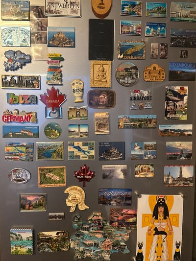 A metallic gray refrigerator door in a residence in Sint-Niklaas, Belgium, is densely covered with an extensive collection of diverse souvenir magnets and a few additional items.
The magnets, varying in size, shape, and material, include:
* **Location-specific text:** "ANTWERPEN," "PISA," "BARCELONA," "IBIZA," "GERMANY," "CANADA" (including a red maple leaf bottle opener), "MONTREAL," "LUXEMBOURG," "THAILAND," and a large map-shaped magnet of "BULGARIA."
* **Landmarks and scenery:** Depictions of Mont Saint-Michel, Vianden Castle, various cityscapes (e.g., Kalabaka, Arbanasi, Vlissingen), landscapes with lakes and mountains, and historical structures.
* **Figurative and cultural items:** A golden seated Buddha, a cream-colored vase with blue floral patterns, a golden Roman-style helmet.
* **Maps:** A magnet depicting a map of Switzerland.
Other items affixed to the refrigerator include:
* A circular wooden object at the top center.
* A black rectangular smart screen device (e.g., a smart fridge display) positioned on the upper right side.
* A white paper drawing at the bottom right, depicting an Egyptian deity resembling Bastet in golden and white attire, accompanied by three black cats, secured by two small silver magnets.
The items are arranged in a tightly packed, eclectic manner, covering most of the visible refrigerator surface. The scene is evenly lit. No individuals are present.