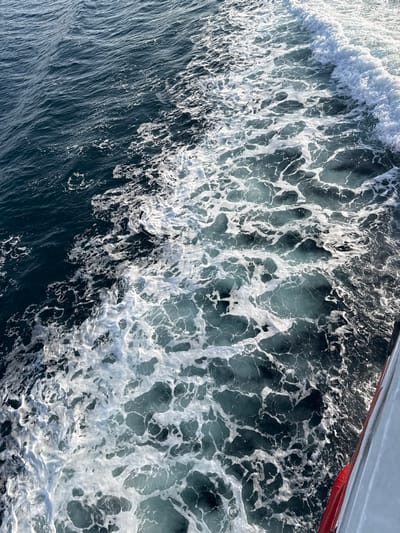 The image displays a close-up, high-angle view of the turbulent wake created by a moving vessel on deep blue-green water near Kamariotissa, Greece. Extensive white foam and aerated water patterns, characteristic of a vessel's propulsion, dominate the central and left portions of the frame. The foam exhibits irregular, frothy formations against the deeper hues of the undisturbed water. On the far right, a section of the vessel's hull is visible; it is light gray or white, featuring a distinct red horizontal stripe along its lower edge. No individuals or other specific objects are discernable within the scene. The primary action depicted is the disturbance of the water surface by the forward motion of a boat or ship.