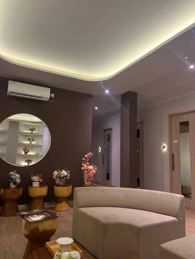 The image depicts a well-lit, modern waiting area, likely in Dumai City, Indonesia. The room features a curved, beige sofa in the foreground, positioned on a light wooden floor. To the left of the sofa, there are three wooden stools, each adorned with a vase of flowers. A round mirror hangs on a brown wall behind these stools, reflecting recessed shelves also decorated with floral arrangements. An air conditioning unit is visible mounted on the brown wall above the mirror. Further into the room, a large, dark pillar stands prominently, and several wooden doors with frosted glass panels are visible. Wall-mounted lights provide additional illumination. The overall ambiance suggests a clean and tranquil space, possibly a spa, salon, or a high-end establishment.