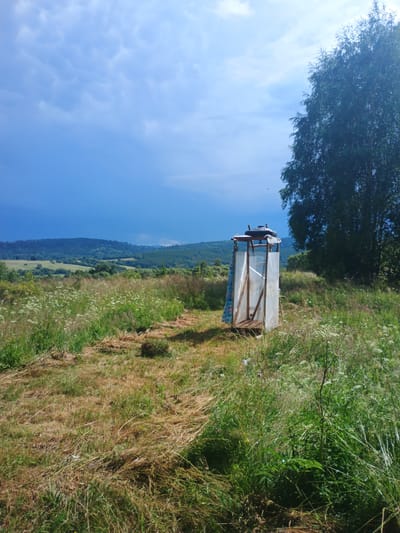 The image depicts an outdoor improvised shower stall set in a grassy field with rolling hills and trees in the background, under a partly cloudy sky. The stall is constructed from a wooden frame and covered with transparent plastic sheeting, with a checkered shower curtain on one side. A black water tank is visible on top of the stall. The surrounding environment is characterized by dry and green grass, wildflowers, and a large tree to the right of the stall. In the distance, lush green hills and a darker, possibly stormy, sky can be seen. The scene appears to be located in Verkhnia Yablunka, Ukraine, likely a rural area. No people or animals are visible in the image.