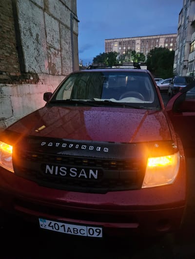 A red Nissan Pathfinder is parked in front of a brick building with a textured concrete facade. The car's headlights and turn signals are illuminated, and its hood features a dark-colored bug deflector with the word "PATHFINDER" in white lettering. The vehicle's license plate is visible, displaying "KZ 401ABC09". In the background, a tall, multi-story apartment building with many illuminated windows can be seen, along with trees and other parked vehicles. The sky is overcast, suggesting either dusk or an early morning. The overall scene depicts an urban environment in Jezqazğan, Kazakhstan.