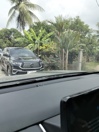 The image captures a frontal view of a black SUV parked outdoors in what appears to be a semi-rural or suburban setting. The vehicle, a Toyota Innova Zenix, is positioned in the foreground, facing the camera. Its license plate, visible as "BL 1498 ZIU," suggests it is registered in Indonesia. Behind the car, a lush green environment unfolds with various trees, including palm trees and banana trees, and some flowering bushes with yellow and pink blossoms. A concrete fence post and a wire fence are also visible in the background, indicating a boundary of some sort. The sky is overcast. The perspective is from inside another vehicle, with a portion of its dashboard and a dark screen visible in the bottom part of the frame. The location is identified as Teupinreudeup, Indonesia.