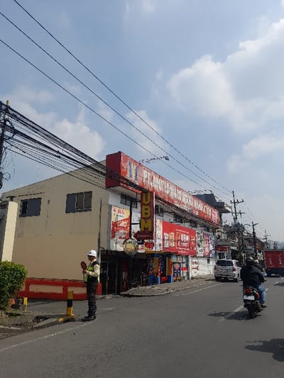 The scene is in front of a large, two-story building with multiple shops and businesses. The building has a large, red and white sign with the words "PT. UNITOMO BERSAMA" visible, which translates to "PT. Unitomo Together". There are also signs with the word "UBM" (possibly an abbreviation for a store name) in bold and other, smaller signs, along with several windows, some open and some closed. A man in a white and blue uniform stands outside, holding a red object. There is a red, yellow, and black barrier on the ground in front of him. The street is paved asphalt with a concrete sidewalk. In the background, a white van is parked to the left, with a red motorcycle to the right. There is a man riding the motorcycle. The sky is blue with white clouds. Power lines run across the sky from the left to the right side of the scene. The location is on a street in the city of Kecamatan Waru, Indonesia, based on the coordinates -7.354 latitude and 112.729 longitude.