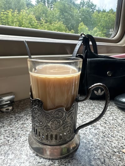 The image captures a tranquil moment within what appears to be a train car, seen from a passenger's perspective. The central focus is a clear glass filled with a light, milky beverage, likely coffee or tea with milk, held firmly in an ornate, silver-colored *podstakannik* (traditional Russian tea glass holder). A small spoon rests inside the glass. The intricate design of the *podstakannik* features swirling floral patterns and a central emblem.
Behind the glass, a black bag, possibly a handbag or small satchel, is visible. A prominent silver Adidas trefoil logo is embossed on the bag. The surface beneath the glass and bag is a speckled grey material, resembling granite or a similar durable countertop.
Through the window to the left, a vibrant green landscape with numerous trees under a bright sky suggests a sunny daytime journey, likely during spring or summer. The window frame and the general interior aesthetic are consistent with a train compartment.
The scene evokes a sense of calm and travel, characteristic of long-distance train journeys, where such glass holders are common, especially in Russia, aligning with the provided location context of Куркачи, Russia. No people are visible, but the presence of personal items implies a passenger enjoying the journey.