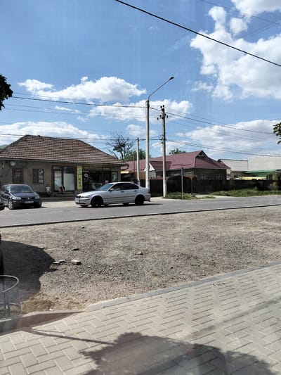 This image depicts a street scene in Congaz, Moldova, on a sunny day with scattered clouds. The foreground shows a paved walkway with a curb separating it from a gravel area. A metal trash receptacle is visible on the left. The main subject of the image is a two-lane asphalt road.
On the left side of the road, a dark-colored hatchback car with license plate Vvh 914 is parked in front of a building with a tiled roof. Two people are sitting on the steps of this building. The building appears to be a commercial establishment with storefront windows displaying various items. Visible text on the storefront includes "ШИНОМОНТАЖ" (tire fitting) and other Cyrillic words, suggesting a tire service or auto repair shop. A sign with "САЛОН МОДНЫЕ" (fashion salon) is also visible.
On the right side of the road, a silver sedan is driving past. Across the street, several residential houses with tiled roofs are visible behind a dark fence. A lamppost and utility poles with power lines are prominent features in the midground. The overall impression is a typical day in a small town or village. The lighting suggests it is daytime, likely late morning or afternoon.
