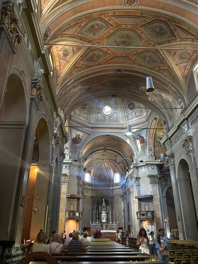 The image shows the interior of the Church of San Vittore in Locarno, Switzerland. The high vaulted ceiling is intricately decorated with frescoes and geometric patterns in shades of orange, green, and gold. Natural light streams in from windows, illuminating the ornate architecture and casting shadows on the pews. Several people are seated on the wooden pews, facing the altar area, which is visible in the distance. Others are standing and walking within the church. Two women in casual clothing, one wearing a light blue cardigan over a white top and the other a white tank top and white shorts, are walking towards the viewer on the right side of the nave. The church appears to be open to visitors and worshippers, with a serene and reverent atmosphere.