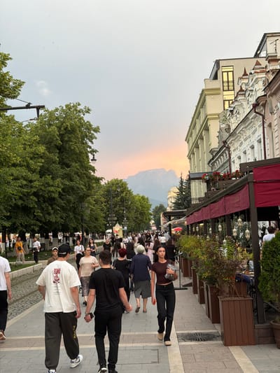 This image depicts a bustling street scene in Verkhniy Alkun, Russia. The street is lined with trees on the left and buildings with awnings and outdoor seating on the right. Numerous people are walking in both directions on the pedestrian walkway. The sky exhibits a soft sunset glow with visible mountains in the distance. People are dressed in casual summer attire. The general atmosphere suggests a lively urban environment, possibly a commercial or tourist area.