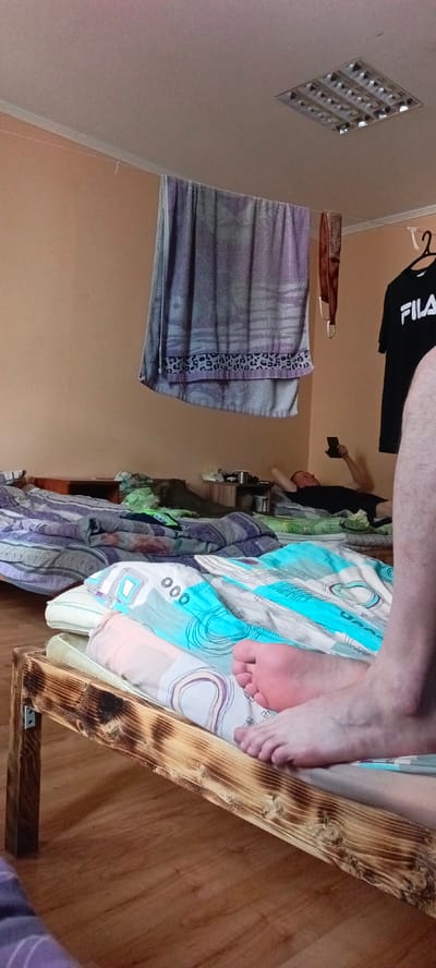 The image depicts a dormitory-style room, likely in Yagodnoye, Russia, characterized by multiple beds. The room has a light-colored ceiling with a fluorescent light fixture. A large towel hangs from a clothesline strung across the room. Adjacent to it, a brown fabric item is also hanging. A black t-shirt with a white "FILA" logo is hanging on a hanger from a hook on the wall.

In the background, a man is lying on a bed, holding a mobile phone. The beds are covered with various patterned comforters. The floor is made of wooden planks. In the foreground, a person's bare feet are visible, resting on a bed with a blue and white patterned comforter. The bed frame in the foreground is made of rough, burnt wood. The overall impression is one of a utilitarian living space, possibly a temporary accommodation.