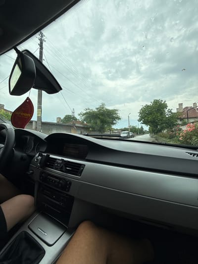 This image captures a first-person view from inside a car looking out through a rain-splattered windshield onto a street in Krushare, Bulgaria. The sky is heavily overcast with dense clouds, suggesting either an impending rainstorm or recent rainfall, further supported by the small water droplets visible on the glass.
Inside the vehicle, the dark grey dashboard and a section of the center console, featuring air vents and climate controls, occupy the right side of the frame. A portion of the black steering wheel is visible towards the bottom right. Hanging from the rearview mirror is a distinct red, leaf-shaped car air freshener with the brand name "Areon" clearly printed on it. The rearview mirror itself is also visible, attached near the top center of the windshield. In the bottom right corner, parts of a person's bare legs are visible, suggesting a driver or passenger is occupying the seat.
Outside the car, the street is lined with lush green trees. Glimpses of houses with reddish-brown tiled roofs can be seen through and above the foliage. A prominent utility pole with numerous overhead wires stands in the mid-ground. Further down the street, a white car is discernible. The scene depicts a typical daytime setting in what appears to be a residential or semi-rural area.