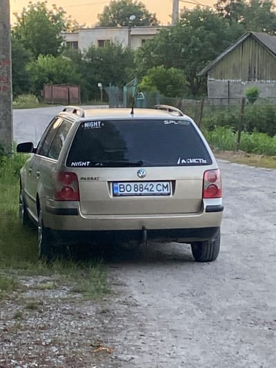 The image shows the rear of a gold Volkswagen Passat station wagon parked on a dirt road in Tovstoluh, Ukraine. The car has several stickers on its rear window. The license plate reads "BO 8842 CM". The surroundings are rural, with trees, houses, and fences visible. The sky is overcast. There are no people visible in the image.