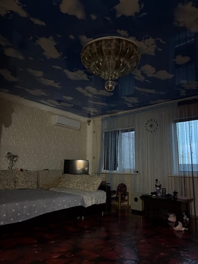 The image depicts a dimly lit living room. The ceiling is a dark blue with a cloudy sky pattern and features a large, ornate chandelier with a disco ball hanging below it. The walls have a textured, circular pattern. On the left side of the room, a sofa is covered with a light blue throw blanket with white stars. An air conditioning unit is mounted on the wall above the sofa. In the center of the room, a television is placed on a surface. To the right, there are several windows covered by sheer curtains. A large wall clock with black numbers is visible on the wall between two windows. In the foreground, the floor is dark and appears to have a glossy, reflective surface with a reddish-brown hue. On a dark wooden table to the right, various objects are placed, including figurines and what appears to be a jar. A small chair is also visible near the windows. The overall impression is of a residential interior.