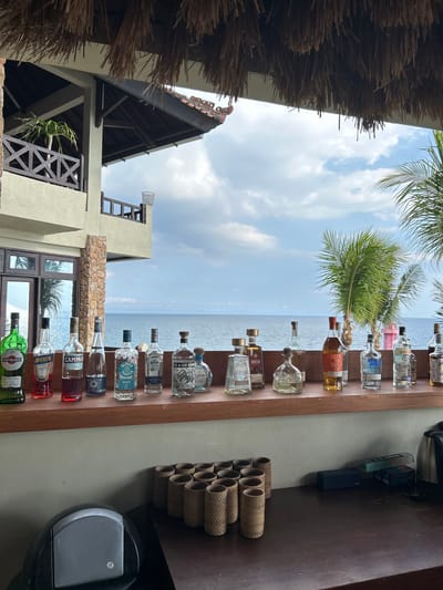 The image depicts a bar area in Senggigi, Indonesia, with a view of the ocean and a building. In the foreground, a wooden bar counter displays an assortment of liquor bottles, including Martini & Rossi, Aperol, Campari, several tequila bottles (including 1800 Tequila, Corralejo Tequila, and Don Julio), Glenmorangie, Absolut Vodka, and Gordon's Gin. In front of the bar counter, there are several woven cylindrical holders and what appears to be a coffee machine. The background shows a multi-story building with wooden balconies and a thatched roof structure extending from the foreground. Beyond the building, the ocean is visible under a partly cloudy sky, with palm trees framing the right side of the image. No people are present in the scene.