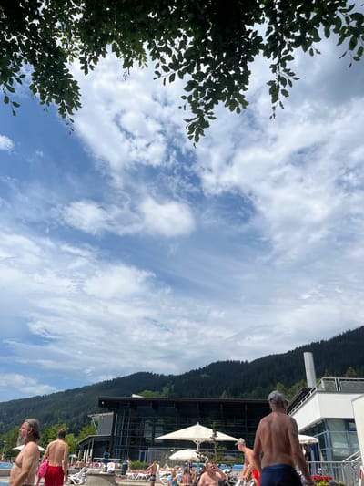 The image depicts an outdoor swimming pool area on a cloudy day, likely in Oberammergau, Germany, given the mountainous backdrop. In the foreground, several shirtless men are visible. One man on the far left, with a long grey beard, is looking to his left. Next to him, another man in red swim shorts is facing away from the camera. A man in a grey baseball cap and blue swim trunks is in the middle-right of the frame, also facing away. Behind them, other swimmers and people relaxing on lounge chairs are present, some under umbrellas. A modern building with large glass windows is visible in the midground, situated against a densely forested mountain range. Overhead, dark green tree branches with leaves frame the top of the image, with a bright blue sky with scattered white clouds above.