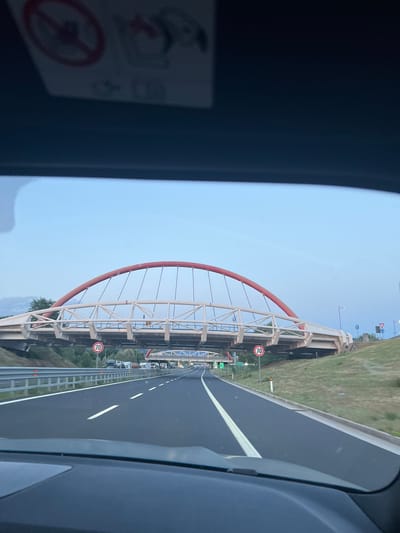 A view from inside a vehicle traveling on a highway approaching a modern, white and red arched pedestrian bridge. The bridge spans across the highway, with traffic lanes visible beneath it. A circular red sign with the number "70" is posted on the right side of the highway, indicating a speed limit. The sky is a clear, pale blue, suggesting either early morning or late evening. The highway is paved with asphalt and has white lane markings. The surrounding landscape includes grassy slopes and some trees. The image is taken from the driver's perspective, looking forward through the windshield. The dashboard of the vehicle is partially visible in the foreground. The specific location in Gorizia, Italy, is not determinable from the visual information provided in the image.