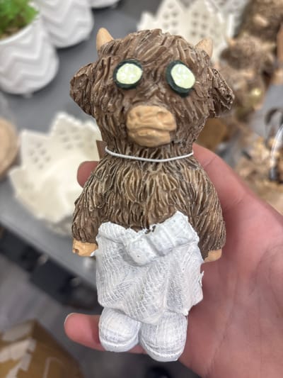 A person is holding a small, brown, figurine of a goat with two light green circular eyes outlined in dark green. The goat has a textured fur-like appearance and a light brown snout and ears. It is wearing a white garment that resembles a dress or apron, with a ruffled hem. The figurine is being held in a person's hand, with their fingers visible. The background is blurred, showing what appears to be a retail setting with shelves and other decorative items. There is no visible text.