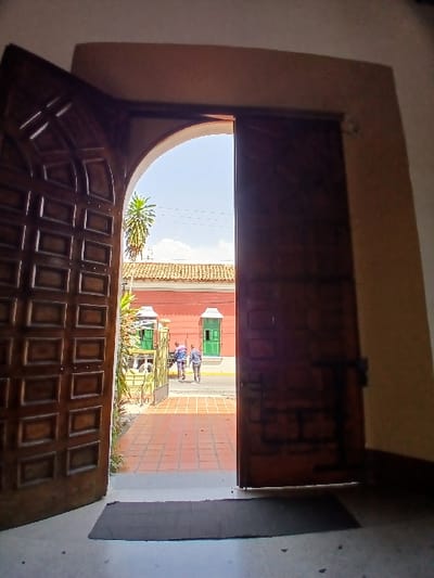 The image depicts a forensic view from inside a building looking out through a large, open, arched wooden doorway in the city of San Joaquín, Venezuela. The foreground shows a dark rectangular floor mat on a light-colored floor, transitioning to reddish-brown square tiles that extend outside. The doorway consists of two dark brown wooden panels, each with multiple recessed rectangular patterns, held open within a light-colored archway.
Beyond the doorway, the scene is brightly lit by sunlight. The exterior features a paved area leading to a street. Across the street, a prominent reddish-orange building with a terracotta-colored roof is visible. This building features at least two distinct green-framed windows or decorative structures with white trim. A tall palm tree is seen against a clear blue sky with minimal white clouds. Green foliage is present on the left side, near the building's exterior.
Two adult individuals are observed in the mid-ground, walking away from the viewpoint on the paved area or street. One individual wears a light-colored shirt and dark pants, while the other wears a patterned shirt and dark pants. No specific actions or interactions between the individuals are discernible.
