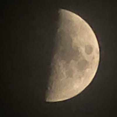 The image shows a close-up view of the moon against a dark, starless sky. Approximately half of the moon is illuminated, displaying craters and surface details. The unilluminated portion is not visible. The image is a photograph taken with a camera, likely through a telescope or with a telephoto lens, given the level of detail of the lunar surface. There are no indications of people, objects, or any urban environment, thus it is impossible to determine any location in Peterborough, United Kingdom.
