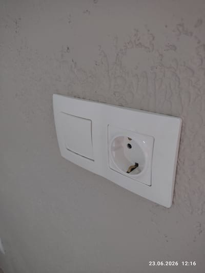 The image shows a white electrical outlet on a textured, off-white ceiling. The outlet has a cover plate and a circular socket with two visible metal contacts and a central grounding pin. Below the socket is a rectangular cover, possibly for another electrical component or a blanking plate. The ceiling texture appears uneven with some darker, irregular patches, suggesting potential water stains or imperfections in the plaster.

The image has a timestamp in the upper right corner that reads "23.06.2026 12:16". This indicates the photograph was taken on June 23, 2026, at 12:16 PM. There are no people visible in the image. The setting appears to be indoors, likely a room in a residential or commercial building, given the presence of the electrical outlet. The lighting suggests it is daytime. The location context provided is Kuyuki, Russia.