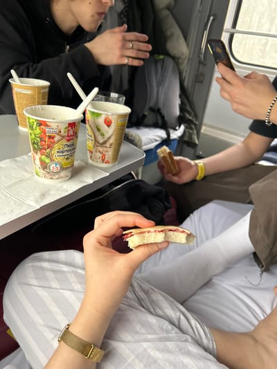 This image depicts the interior of a train compartment in Yosypivka, Ukraine, during daylight hours. Three individuals are visible. In the foreground, a person's hands and lap are shown. This individual holds a partially eaten sandwich, appearing to be white bread with a red filling. A gold-toned mesh band watch is worn on their left wrist, and they are clad in light-colored striped trousers. To the immediate right, another person holds a small, rectangular sandwich-like item in their left hand and a dark-colored smartphone in their right hand, appearing to be in use. This individual wears dark trousers, a yellow wristband on the right wrist, and a black and white beaded bracelet on the right wrist. Further to the left in the midground, a third person's upper torso and hands are visible. This individual wears a dark long-sleeved garment and a silver ring on their left ring finger.
A light-colored table extends across the midground. On the table, two paper cups containing instant mashed potatoes are present, one with a white plastic spoon. The visible cup packaging includes Ukrainian text, "ПЮРЕ КАРТОПЛЯНЕ" (mashed potatoes), and "MADE IN UKRAINE" labeling. An empty, translucent glass and a white paper napkin are also on the table. The background features a train window on the right, showing a bright, undifferentiated outdoor scene. Dark blue upholstery is visible on what appears to be a train seat or bench, and a grey backpack hangs in the background on the left. The scene suggests passengers consuming food and beverages while traveling.