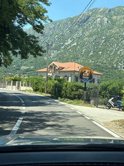The image shows a road curving through a mountainous, green landscape. A sign for "AUTO CAMP Dule" is visible, featuring a stylized caravan, trees, and water. Behind the sign, a building with a terracotta roof and stone facade can be seen. In the background, a steep, rocky mountain covered in sparse vegetation rises against a clear blue sky. Trees line the side of the road, and their shadows stretch across the asphalt. A blue and white scooter is parked on the side of the road near the sign. The scene appears to be set in a rural or semi-rural area, possibly a tourist destination given the campsite sign. The bright sunlight and clear sky suggest it is daytime, likely morning or afternoon, with good weather. The perspective suggests the photo was taken from inside a vehicle, looking out.