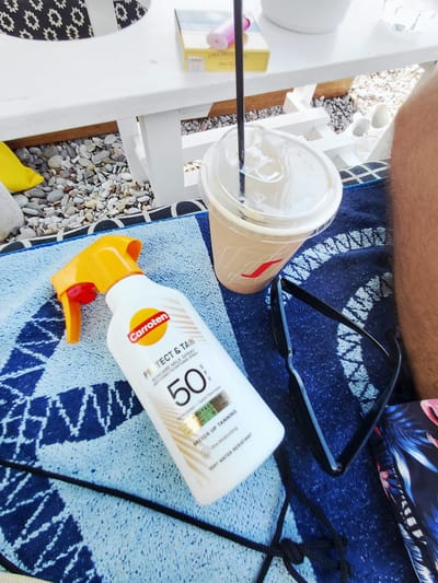 The image captures a close-up view of a beach scene, likely in Municipal Unit of Diakopto, Greece, as suggested by the context. A light blue and white patterned towel lies on the ground, serving as a base for the objects.

On the towel, a white spray bottle of "Carroten Protect & Tan" sunscreen with SPF 50 is prominently displayed. The bottle has a bright yellow spray nozzle and an orange trigger. To the right of the sunscreen, a clear plastic cup with a beige-colored beverage and a straw sits next to a pair of black sunglasses.

In the background, a white table with some items on it, including a yellow packet and a pink object, is visible. The table is placed on a surface covered with small pebbles, typical of a beach environment. A person's leg, with visible hair, is partially in the frame on the right side. A blue and white patterned fabric, possibly part of swimwear or a bag, is also visible in the lower right corner.