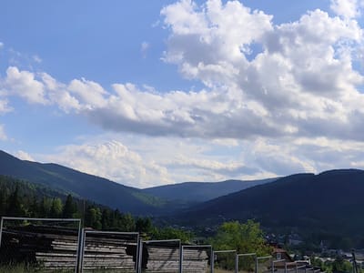 This is a landscape photograph taken in Mykulychyn, Ukraine. The scene depicts a view of rolling, forested mountains under a bright blue sky dotted with fluffy white clouds. The mountains are layered, with the closest ones appearing a deep green and the further ones fading into a dusky blue, suggesting depth and distance. In the foreground, there are fences made of chain-link and wooden planks, some of which are stacked with what appear to be building materials or lumber.
To the right of the frame, the lower slopes of the mountains transition into a village, with houses and rooftops visible amongst the trees. The presence of houses and the arrangement of lumber in the foreground suggest a rural or semi-rural residential area.
The lighting indicates it is daytime, likely late afternoon or early evening, given the soft shadows and the warm tones in some parts of the sky. The weather appears to be fair and pleasant, with the clouds adding visual interest to the sky. The overall mood is peaceful and serene, capturing the natural beauty of the mountainous region. No text is visible in the image.