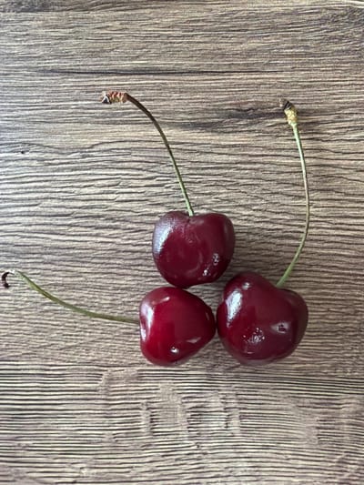 Three dark red cherries with thin green stems are arranged on a wooden surface with horizontal grain. One cherry is positioned at the top center, with its stem arching to the left. Another cherry is located below and to the left of the first, with its stem extending to the lower left. The third cherry is to the right and slightly below the first, with its stem curving upwards to the top right. The wooden surface is textured and appears to be a table or countertop. There are no people or animals present in the image, and no actions or interactions are depicted. The environment is an indoor setting, with artificial lighting. The location of Shashmurka, Russia is not depicted or implied by any elements within the image.