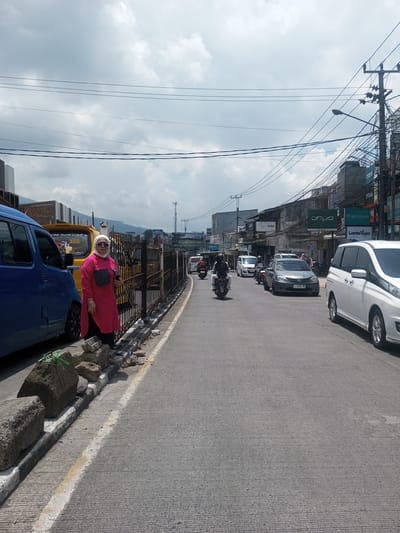 A daytime street scene shows a woman standing on a sidewalk next to a curb adorned with gray rocks. The woman, wearing a pink top, dark pants, and a white head covering, poses for the camera. Behind her, a blue van is parked, along with a yellow vehicle further down the street. A black metal fence runs parallel to the curb.
The street is filled with moving traffic, including motorcycles and various cars. Buildings line the other side of the street, displaying advertisements for products like Oppo and Wedrink, indicating a commercial area. Overhead, a tangle of power lines stretches across the sky, which is partly cloudy.
The location is likely an urban area. The specific city or district information, Xayabury District, Laos, is not evident in the image.
