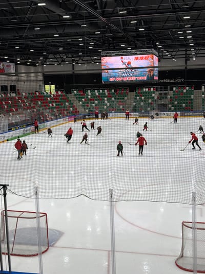 An aerial, wide-angle shot captures an ice hockey rink during what appears to be a training session. The rink is full of young players wearing various colored hockey uniforms, predominantly red, yellow, and black. Several adults, likely coaches, are also present on the ice, some of whom are wearing red jackets. The players are spread across the ice, engaged in drills, with hockey sticks in hand and pucks visible. Two hockey nets are positioned at either end of the rink. The stands, filled with red and green seats, are visible in the background, mostly empty. A large video screen hangs above the rink, displaying graphics and the time "10:19:42". The overall environment is that of an indoor sports arena, with exposed ceiling infrastructure and bright lighting. The location is the Arena Sosnowiec, located in Sosnowiec, Poland, a multi-purpose indoor arena.