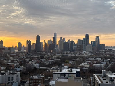 The image shows a cityscape in Jersey City, United States of America, with a skyline of skyscrapers visible in the distance. The sky is overcast with clouds, and the sun is setting, casting an orange glow on the buildings. The foreground contains residential and commercial buildings, with some trees and roads in between. There are no people visible in the image.