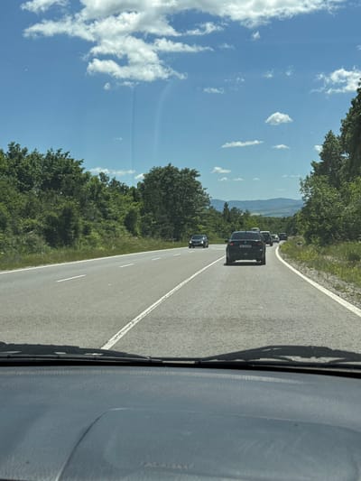 The image captures a view from inside a vehicle driving on a two-lane asphalt road through a rural, forested area. The road has white lane markings. Several cars are visible on the road ahead, traveling in the same direction. The surrounding environment is characterized by lush green trees and vegetation lining the sides of the road, with rolling hills and mountains visible in the distance under a bright blue sky with scattered white clouds. The scene suggests a sunny day. The location is identified as Ponor, Bulgaria.