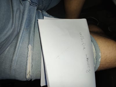 The image shows a close-up view of a person's lap area, featuring denim jeans with a distressed section. A white sheet of paper is held across the lap, with handwritten text in black ink visible on its surface. The text reads "collective memory." The environment appears to be indoors, with dark shadows suggesting a dimly lit setting. No specific location in Eutin, Germany, can be identified from the visual information provided in the image.