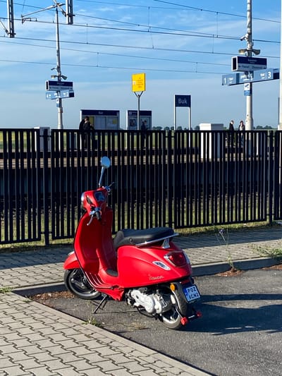 A red Vespa scooter is parked on a paved area next to a black metal fence. The scooter is facing away from the camera, with its rear wheel and license plate clearly visible. The license plate reads "POZ 6CN2". Behind the fence, a train platform is visible with several signs and poles. One sign indicates "Buk" as a destination. A few people are standing on the platform, some appearing to be waiting for a train. The sky is blue with some clouds, and the sun is shining, casting shadows on the ground. The scene appears to be a train station in or near Dobieżyn, Poland.
