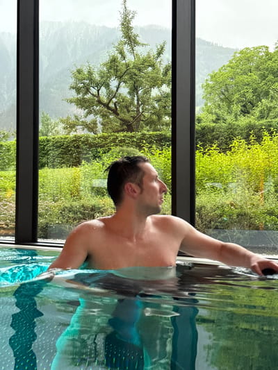 The image shows a shirtless man in a swimming pool. He is looking to his right, with his left arm resting on the edge of the pool. The pool has turquoise water with reflections. Behind the pool is a large window overlooking a lush green landscape with trees and mountains in the distance, possibly suggesting a location in or near Mayrhofen, Austria, given the mountainous terrain. The atmosphere appears to be overcast or misty, contributing to a soft, diffused light.