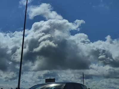 In Svētciems, Latvia, a dark gray cloud dominates the sky, casting a shadow over the landscape. A tall, dark pole, possibly a light pole, stands prominently in the foreground, bisecting the frame vertically. Below the pole, the roof and windshield of a dark-colored vehicle are visible, reflecting the bright sunlight. The vehicle appears to be parked or moving slowly. In the background, power lines stretch across the frame, supported by wooden poles, indicating a more developed area. The sky is a mix of deep blue and patchy white clouds, suggesting a dynamic weather system.