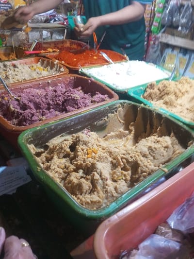 Various colorful dips fill plastic containers at a market stall. A person is preparing ingredients near a red chili paste. Dips range from white, purple, brown, and tan. Spoons are placed within each dip, ready for serving. Packaging of goods hangs nearby.