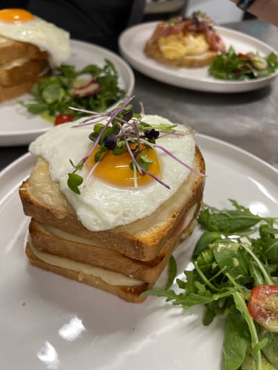 A close-up, eye-level shot captures a stack of three toasted bread slices, each layered with melted cheese and a thin slice of ham, topped with a fried egg with a runny yolk. The sandwich is garnished with delicate purple sprouts. To the right of the sandwich, a small side salad consists of arugula, a halved cherry tomato, and a light green dressing. The dish is presented on a white plate, which rests on a grey countertop or table. In the background, out of focus, are two other plates of food, one with a similar sandwich and salad, and another with scrambled eggs and bacon. The overall scene suggests a breakfast or brunch setting. There is no visual information to determine the location in the city of Vysoké Tatry, Slovakia.