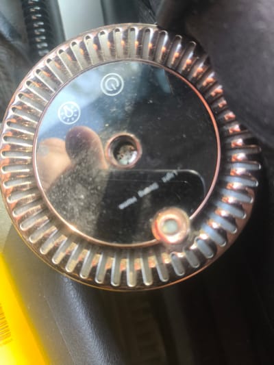 A top-down close-up shot shows a rose gold colored humidifier with a ribbed edge. The humidifier has a glossy black top with a power button symbol, a mist symbol, and a small, circular opening with a metal grate inside. There is also a small, circular, rose gold button located near the bottom right of the humidifier. The humidifier is resting on a dark, textured surface. A yellow object is partially visible in the bottom left corner of the image. The image is sharp and well-lit. The humidifier appears to be a portable electronic device. There are no visible signs of the city of Abay, Kazakhstan in the image.