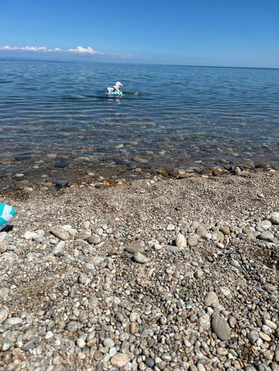 A person in a white hat and striped swimwear floats on a circular inflatable ring in clear, calm water. The shoreline, composed of small, smooth pebbles, slopes gently into the water. In the foreground, a portion of a blue and white patterned inflatable object rests on the pebbles. The sky above is a clear, bright blue with a few wispy white clouds on the horizon. The location is the shore of Lake Baikal in the village of Tolbazikha, Russia.