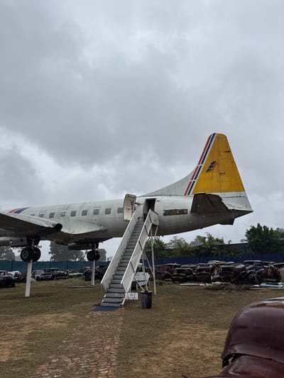 The image shows the tail section and fuselage of a decommissioned passenger airplane, specifically a Convair 580, in a scrapyard or open-air museum. The tail is painted yellow with red, white, and blue stripes. A set of stairs is attached to the aircraft's door, allowing access inside. The environment includes grassy terrain, scrap metal, and the sky is overcast. The location is likely in Kraaifontein, South Africa, where such displays can be found. There is no evidence of any people in the image.