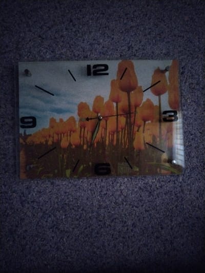 A rectangular wall clock with a printed image of yellow tulips against a cloudy sky is mounted on a textured, dark blue surface, likely a carpet. The clock face displays Roman numerals for 12, 3, 6, and 9, with black hour and minute hands pointing to approximately 1:30. The clock is secured to the surface by two visible silver screws. The lighting is dim, casting shadows on the lower portion of the image and creating a slight glare on the clock face. The scene is indoors. The location of Vaziyo, Uzbekistan is not depicted in this image.