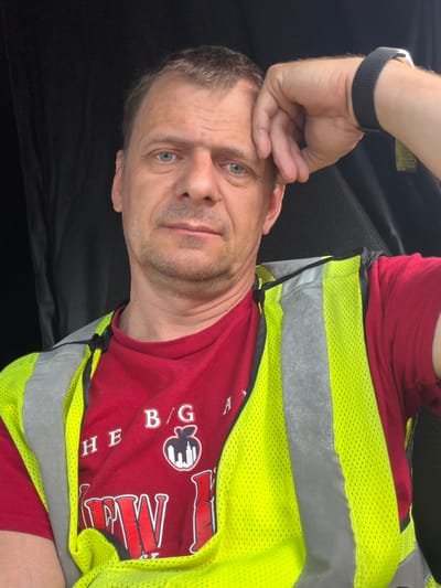 A man lies on his side, his head propped up by his hand. He is wearing a red t-shirt with white text and graphics, and a bright yellow safety vest with gray reflective stripes. The background is dark and appears to be an interior space, possibly a car seat. The man has light blue eyes and short, light brown hair. He has a slightly stubbled jawline and his expression is neutral. The lighting suggests it might be daytime, but the indoor setting makes it difficult to determine the exact time or weather. There is some visible text on his t-shirt, though it is partially obscured by the safety vest. Some legible letters include "H E B / G" and a partial word that appears to be "W V". There is also a graphic of a stylized building or landmark. The image is oriented sideways.
