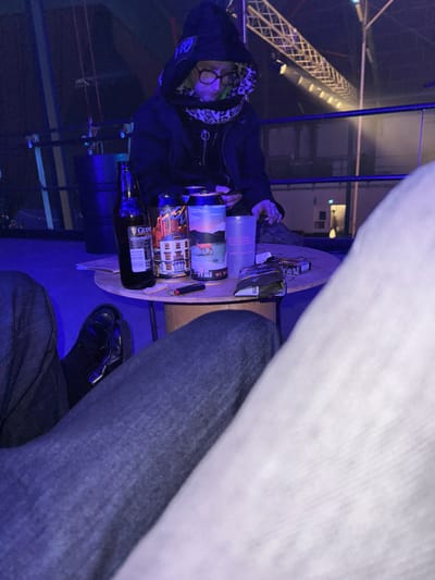 A slanted view depicts a partially visible person wearing a hooded sweatshirt and glasses, seated near a small round table. The table holds an assortment of items including a bottle of Guinness, several cans decorated with vibrant artwork, a plastic bottle, a cigarette pack, and a lighter. Jeans and black shoes are visible in the foreground. In the background, a structure with exposed metal beams and blue lighting creates an atmospheric backdrop.